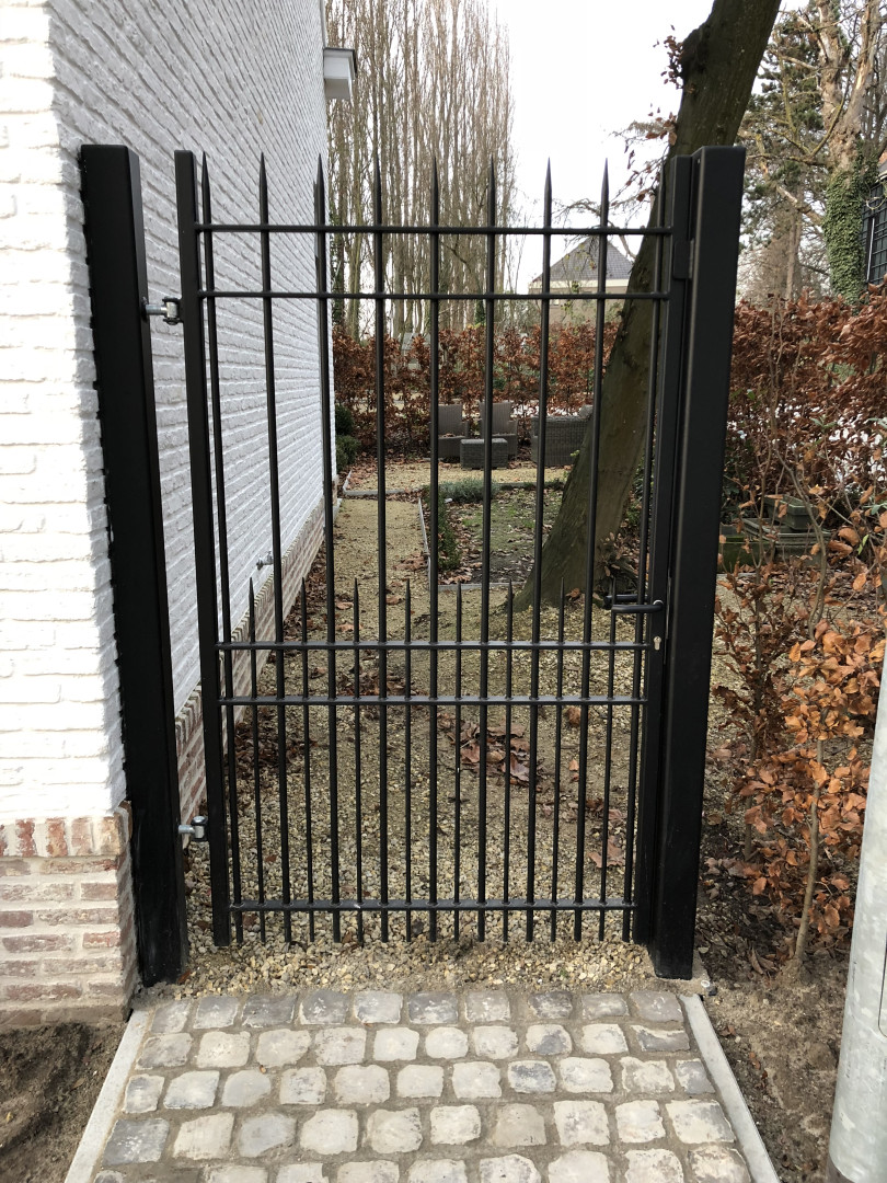 Strakke tuinpoort in zwart gepoederlakt staal met speerpunten Zwarte stalen tuinpoort met verticale spijlen en speerpunten, geplaatst naast een wit bakstenen huis, op maat gemaakt door Laswerken Bellon in Sint-Niklaas.