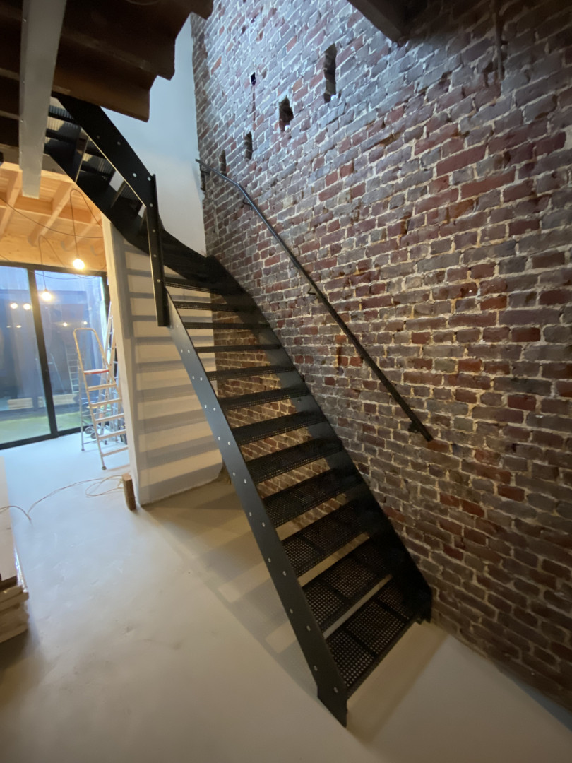 Industriële stalen trap op maat met geperforeerde treden Zwarte stalen trap met geperforeerde treden en strakke leuning, gemonteerd tegen een bakstenen muur in een modern-industriële woning in Sint-Niklaas.