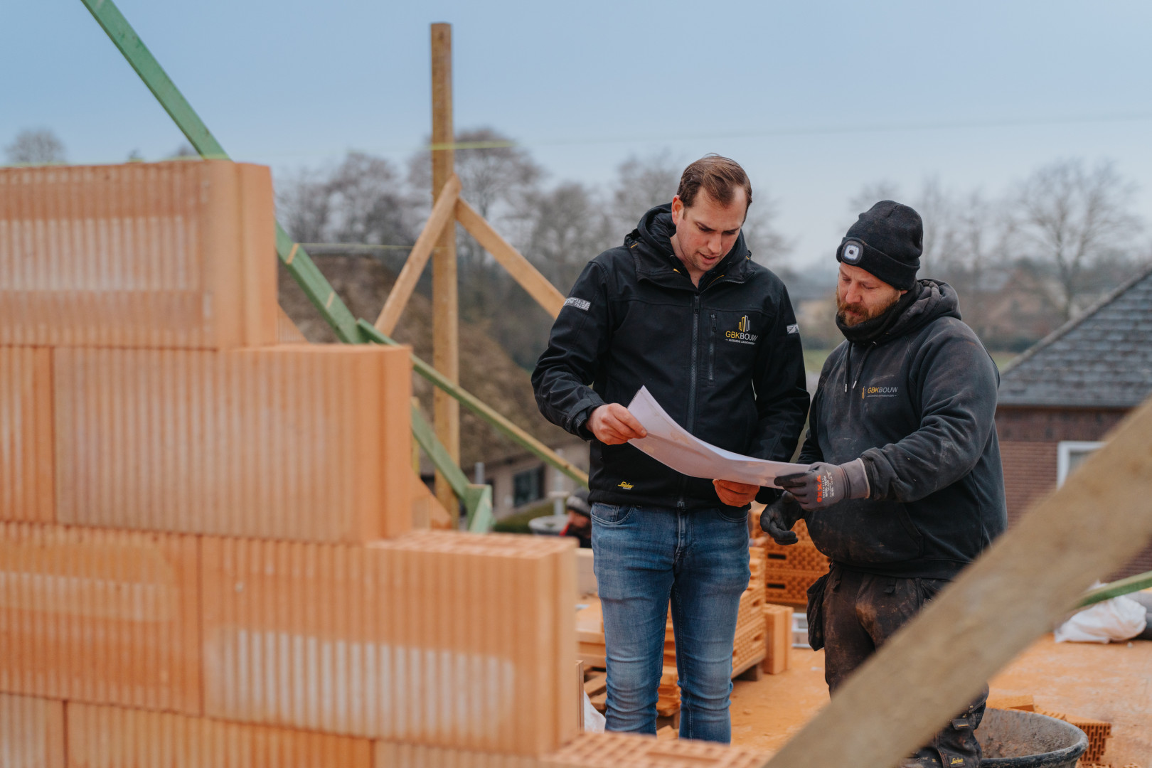 Ruwbouw in uitvoering: planning en opvolging op de werf door GBK Bouw Twee medewerkers van GBK Bouw bespreken bouwplannen tijdens de ruwbouwfase op de werf, omringd door snelbouwstenen en houten constructie-elementen.