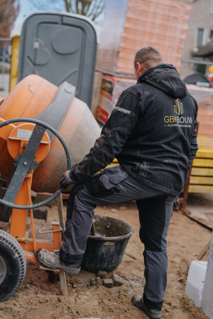 GBK Bouw aan het werk op de werf: voorbereiding van beton Mederwerker van GBK Bouw bedient een betonmolen op een bouwwerf, met werfkledij en zichtbaar logo op de rug, omringd door bouwmaterialen.