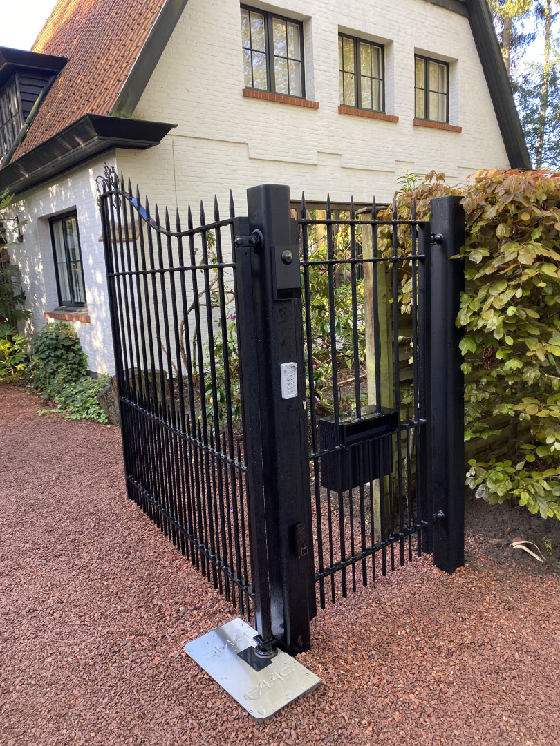 Zwarte smeedijzeren tuinpoort met sierpunten en krullen, geplaatst bij een landelijke woning met witte gevel en rode oprit in Sint-Niklaas.