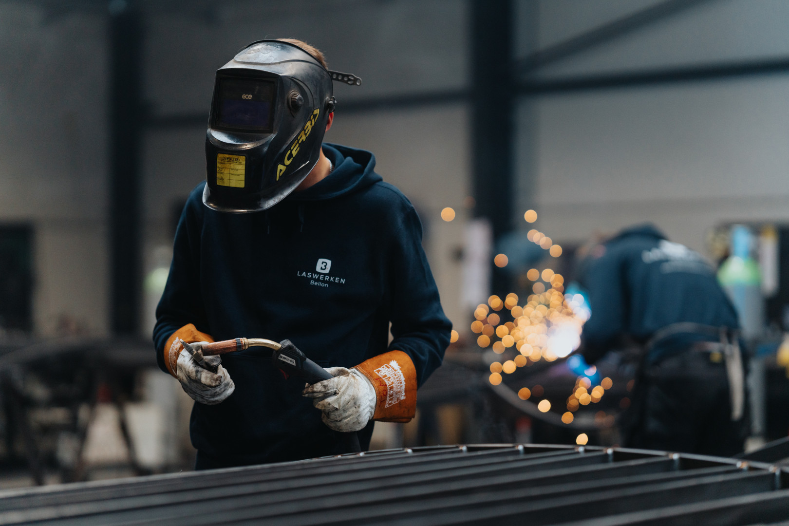 Een lasser van Laswerken Bellon last met precisie een metalen constructie in het atelier, met op de achtergrond vonken en vakmensen aan het werk.