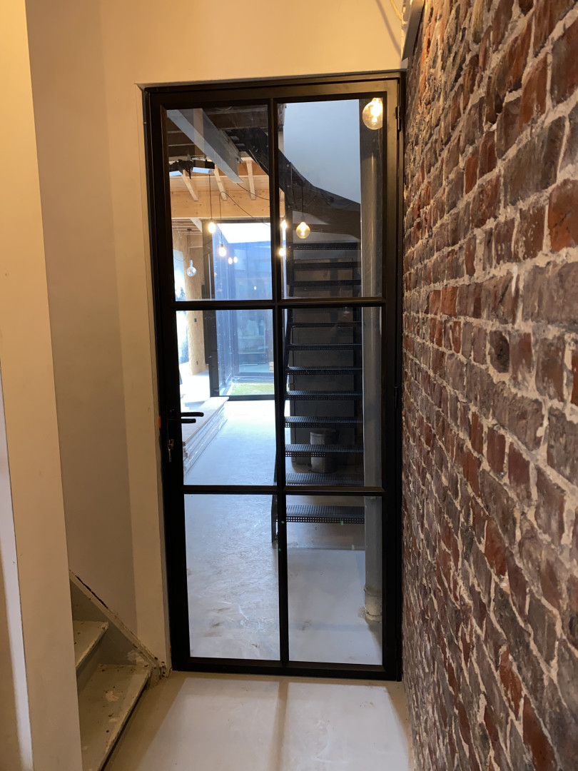 Op maat gemaakte stalen binnendeur met glas, geplaatst in een industri&euml;le woning in Sint-Niklaas, met zicht op trap en houten plafondbalken.