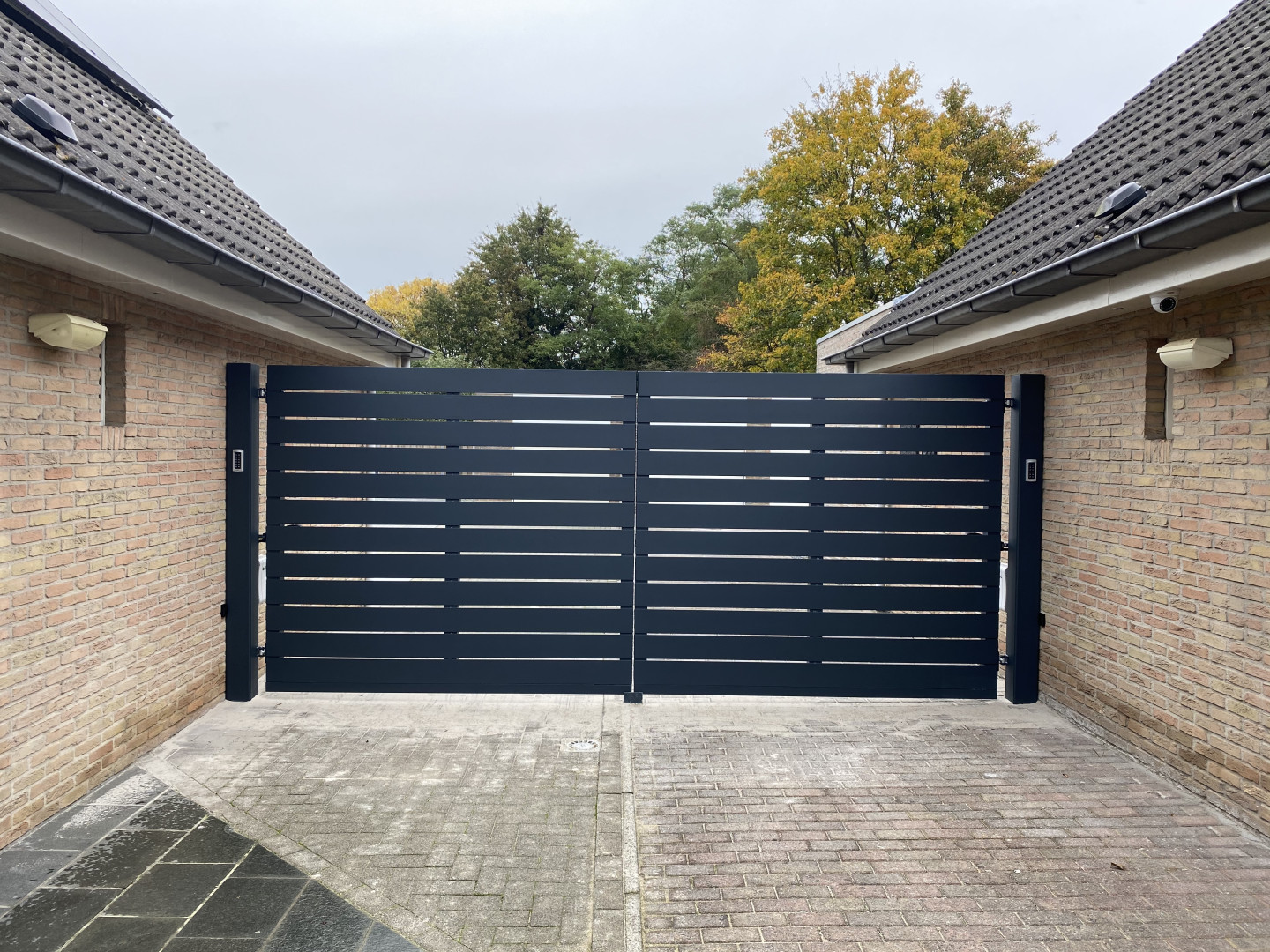 Donkergrijze aluminium poort met horizontale lamellen en ge&iuml;ntegreerde videofonie, geplaatst tussen twee woningen met rode gevelsteen.