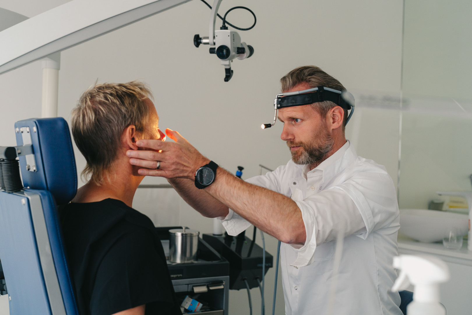 Dermatologisch onderzoek van huidletsel bij The Face Clinic Arts voert een gericht huidonderzoek uit bij een patiënt met gespecialiseerde verlichting in de behandelruimte van The Face Clinic in Lommel.