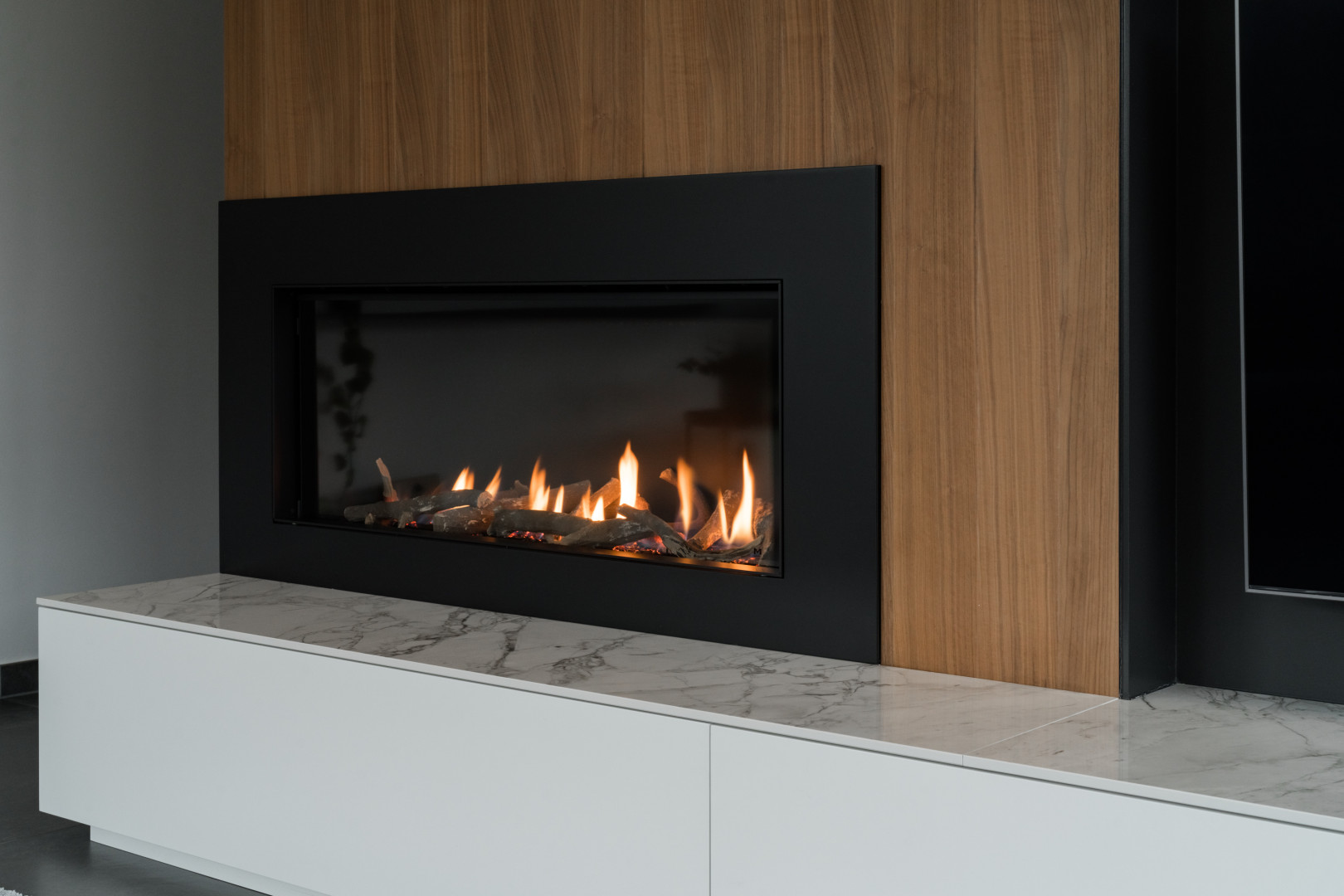 Strakke inbouw gashaard met marmeren en houten afwerking Moderne gashaard ingebouwd in een wand met houtfineer en marmeren blad, naadloos geïntegreerd in een tv-meubel met zwarte omlijsting.