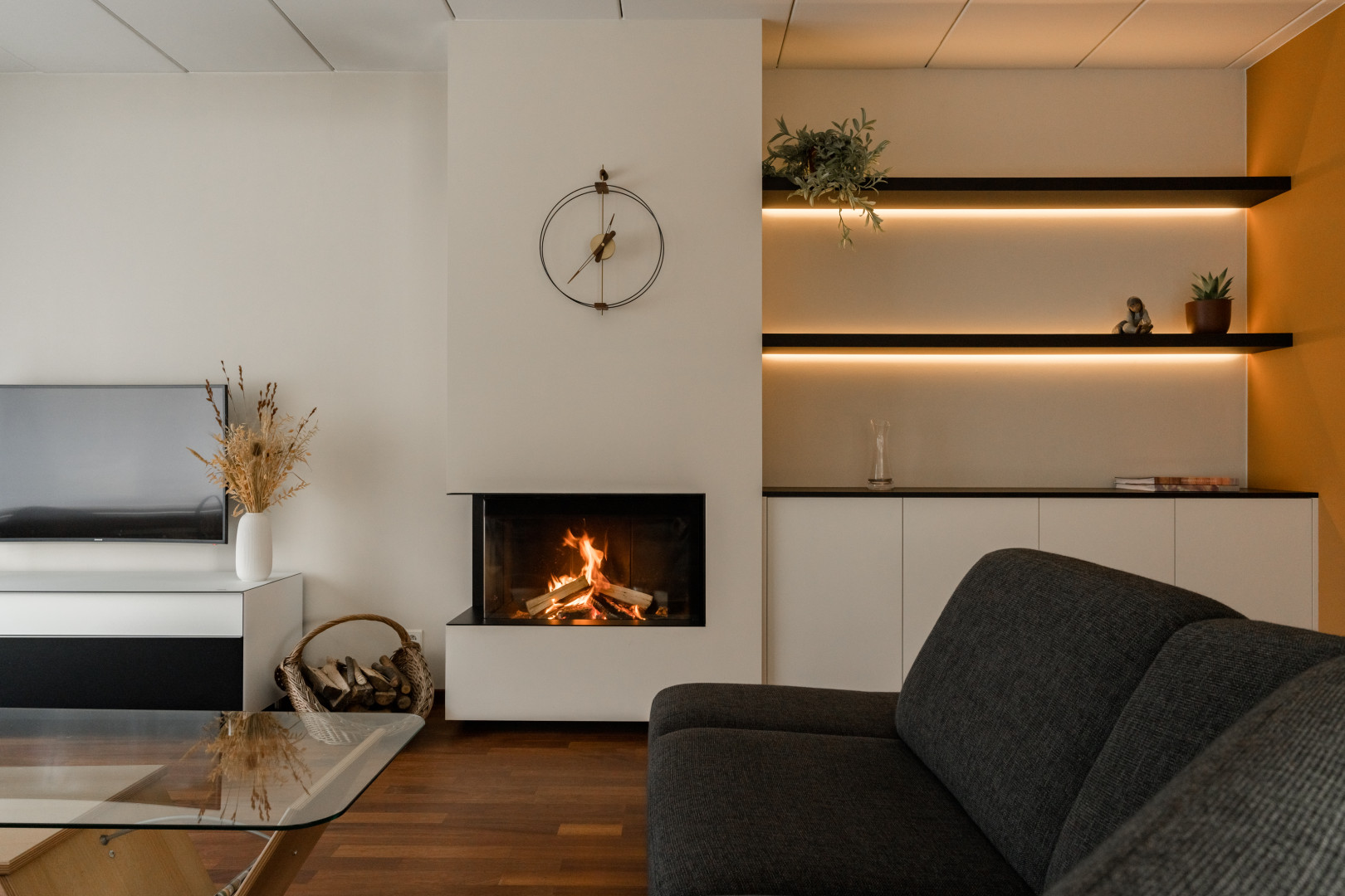 Gezellige hoekhaard met maatwerk wandkast en sfeerverlichting Moderne hoekhaard in witte omkasting, gecombineerd met een maatkast en zwevende legplanken met geïntegreerde ledverlichting in een warme leefruimte.